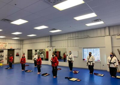 A group of people in martial arts uniforms and belts stand in a spacious, bright training room with blue mats on the floor. Some wear red tops and others wear white, and a few are wearing face masks. Equipment and windows are visible in the background.