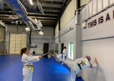 Two people in martial arts uniforms are practicing in a large training room with blue mats on the floor. One holds a target pad while the other executes a high kick towards it. The room has visible ventilation ducts and a partial view of a motivational sign on the wall.