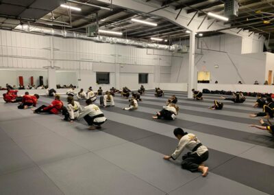 A group of martial arts practitioners, dressed in uniforms with various belts, perform exercises on a large, padded training floor inside a well-lit dojo. The space is industrial with exposed ceiling elements and a white interior.