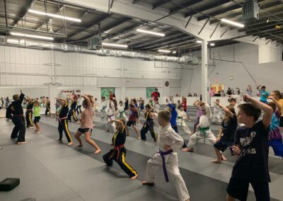 A group of children wearing martial arts uniforms and different colored belts are practicing techniques in a spacious, well-lit dojo. Several instructors are present, assisting and guiding the students. Onlookers and parents watch from the sides of the room.