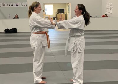 Two women, dressed in white martial arts uniforms, stand facing each other in a dojo with a gray mat. One woman has an orange belt, and the other has a white belt. They are performing a partnered drill, touching palms. A sign in the background reads "LION FORCE STRONG.