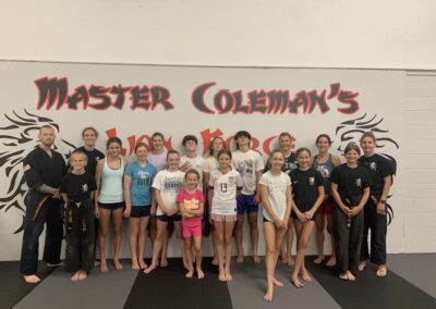 A group of martial artists of various ages, dressed mostly in casual sportswear, pose for a photo in front of a wall with "Master Coleman's" written in large letters. Some wear martial arts belts. The setting appears to be a dojo or martial arts training facility.