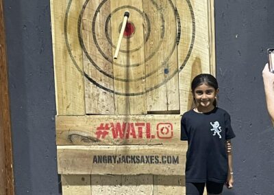 A young girl stands smiling in front of an axe-throwing target, with an axe lodged in the bullseye. The background features a wooden target board with a hashtag "#WATL" in red and the website "angryjacksaxes.com" written on it.