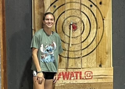 A person wearing a green T-shirt with a cartoon character and black shorts is standing next to an axe-throwing target. The target has an axe embedded in the bullseye. The background and target board feature text, including "ANGRY JACKS" at the top.