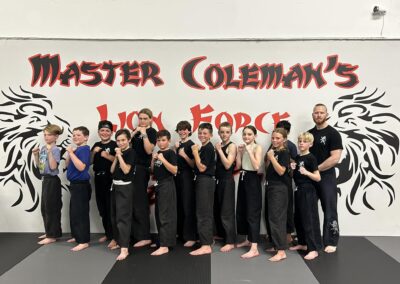 A group of children and a man, all dressed in martial arts uniforms, stand in front of a wall with the text "Master Coleman's Lion Force" and a lion's head motif. The children are posing with their fists raised, ready for karate practice.