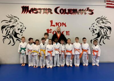 A group photo shows a martial arts class standing in front of a wall with the text "Master Coleman's Lion Force." The instructor, wearing a black and red uniform, stands behind eleven children dressed in white uniforms with colored belts ranging from yellow to green. The US flag is to the right.